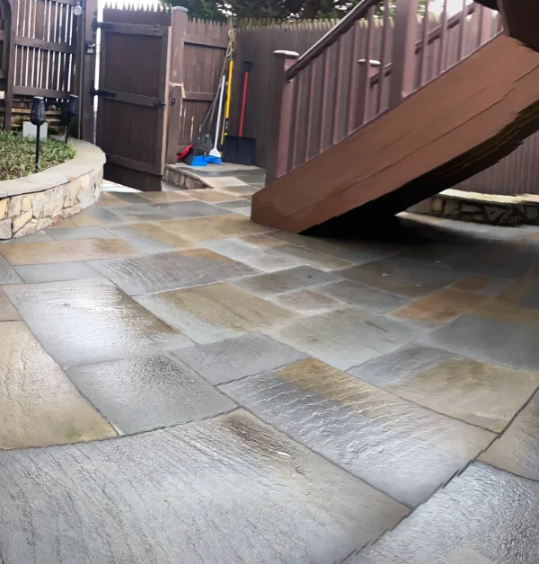 A freshly repaired flagstone patio featuring clean, multi-colored natural stone pavers sits completed next to a wooden deck staircase and a low stone retaining wall leading toward an open back gate.