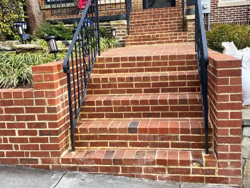 A freshly repointed red brick front porch staircase and matching retaining walls feature clean, uniform mortar joints alongside black wrought iron handrails leading up to a home entrance.