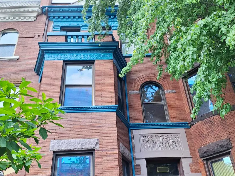 A close-up view of the same historic rowhouse after renovation, showcasing freshly cleaned brickwork and vibrant teal-painted architectural trim framed by lush green trees.