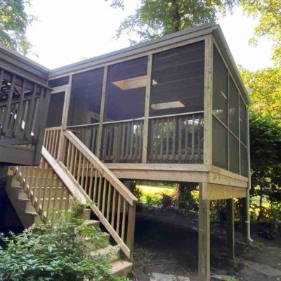 Screened-in porch with wooden railings and stairs, elevated on wooden posts, surrounded by trees.