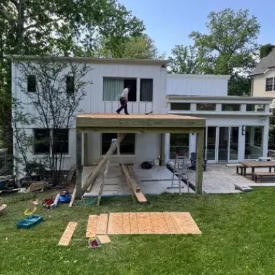 A person stands on the roof of a partially constructed patio at the back of a modern white house.