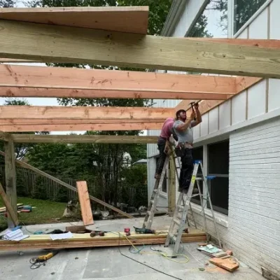 Two workers on ladders constructing a wooden pergola frame attached to a house.