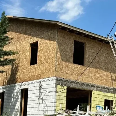 Two-story house under construction with a ladder, surrounded by trees and a fence.