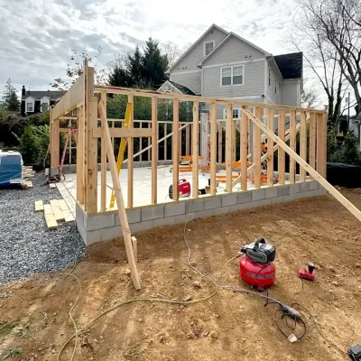 Wooden framing for a building is under construction beside a house on a cloudy day.