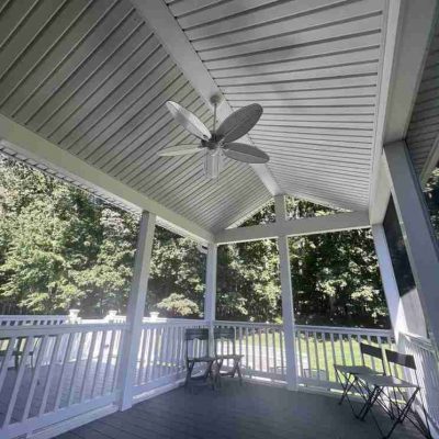 Covered porch with a ceiling fan, white railing, and folding chairs, overlooking a wooded area.